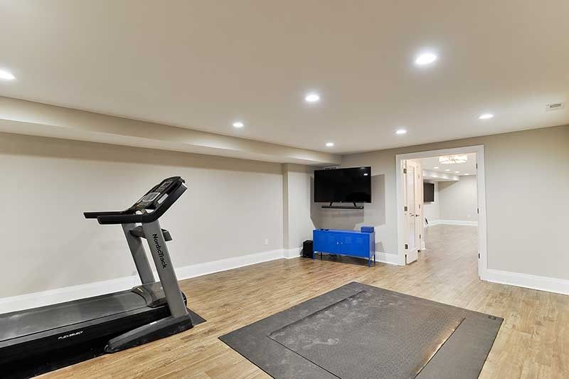 A home gym with a treadmill, a floor exercise mat, a wall-mounted TV, and a blue storage unit on light wood flooring.