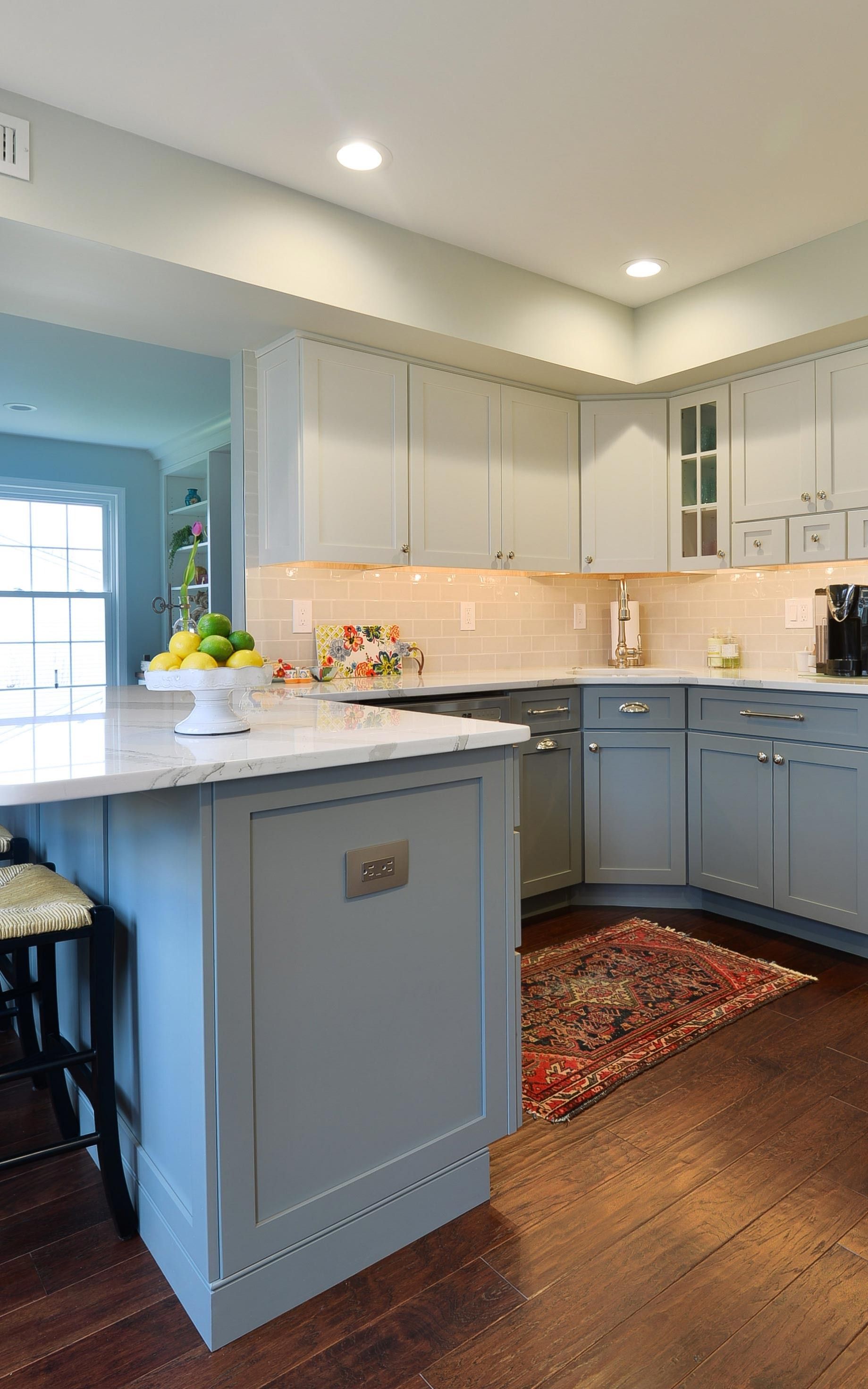 A kitchen features white upper cabinets, light gray lower cabinets, a marble-topped island, and a dark wood floor.