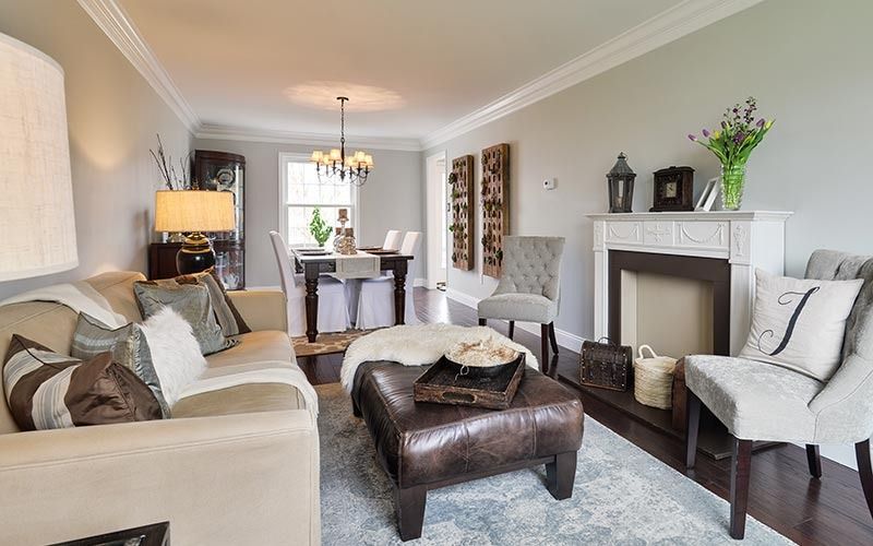 A living room featuring a beige couch, leather ottoman, white fireplace, and a dining area visible in the background.