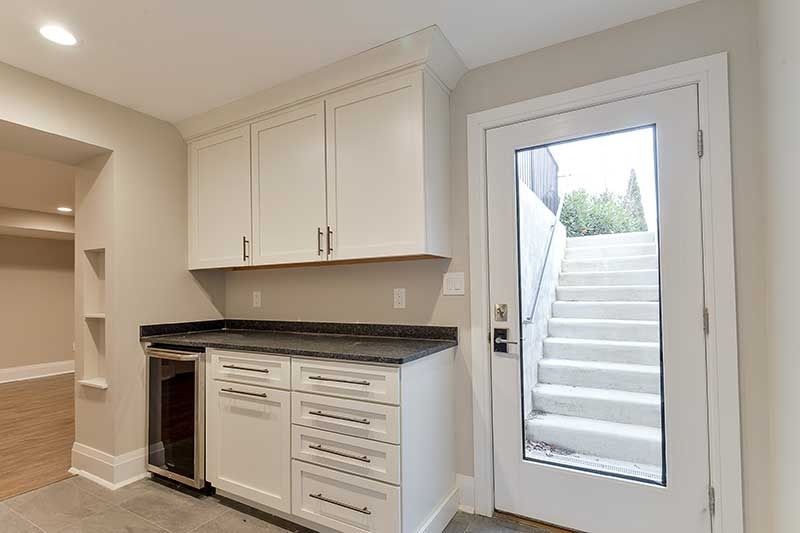 A small wet bar with white cabinets, a dark countertop, and a wine cooler next to a glass door revealing outdoor stairs.