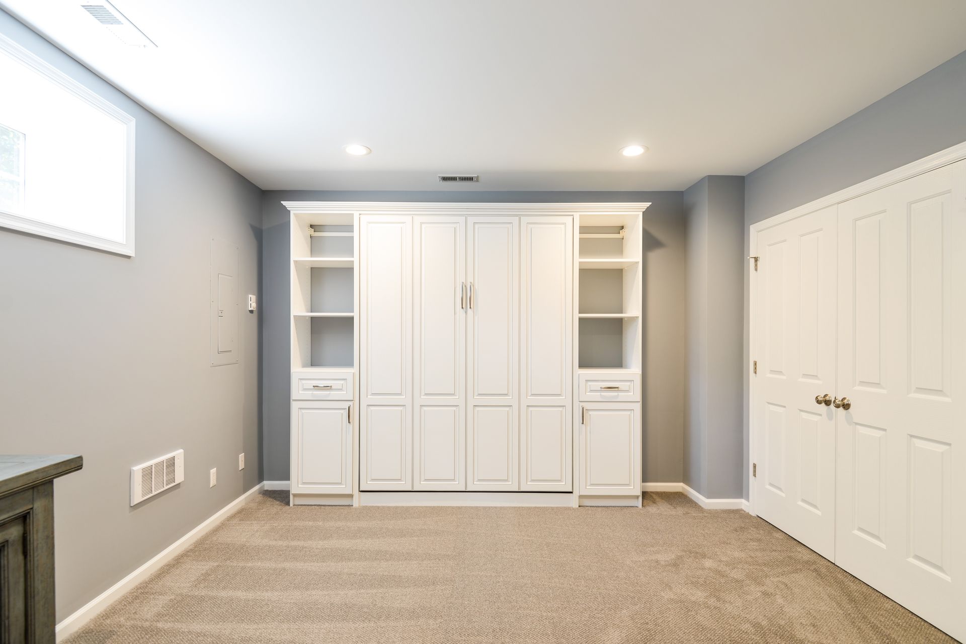 A white Murphy bed with built-in shelving centered against a light gray wall in a carpeted room with white closet doors.
