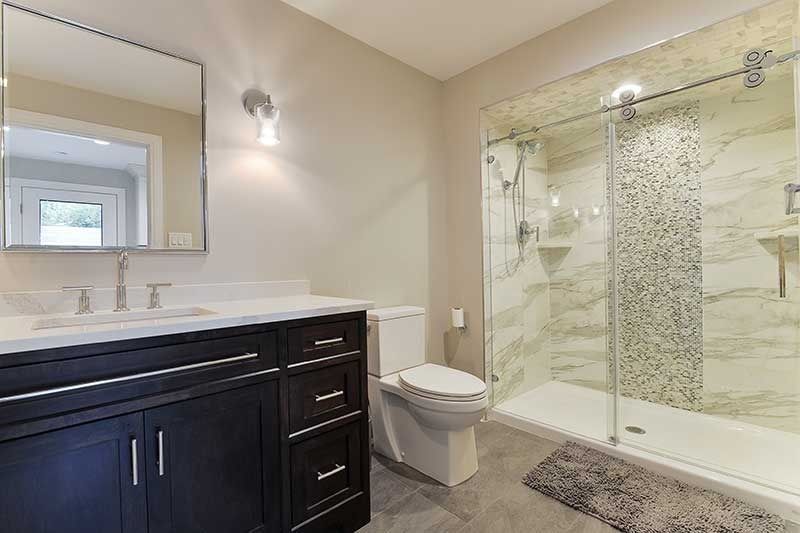 A modern bathroom featuring a dark wood vanity, white countertop, rectangular mirror, toilet, and glass-enclosed shower.