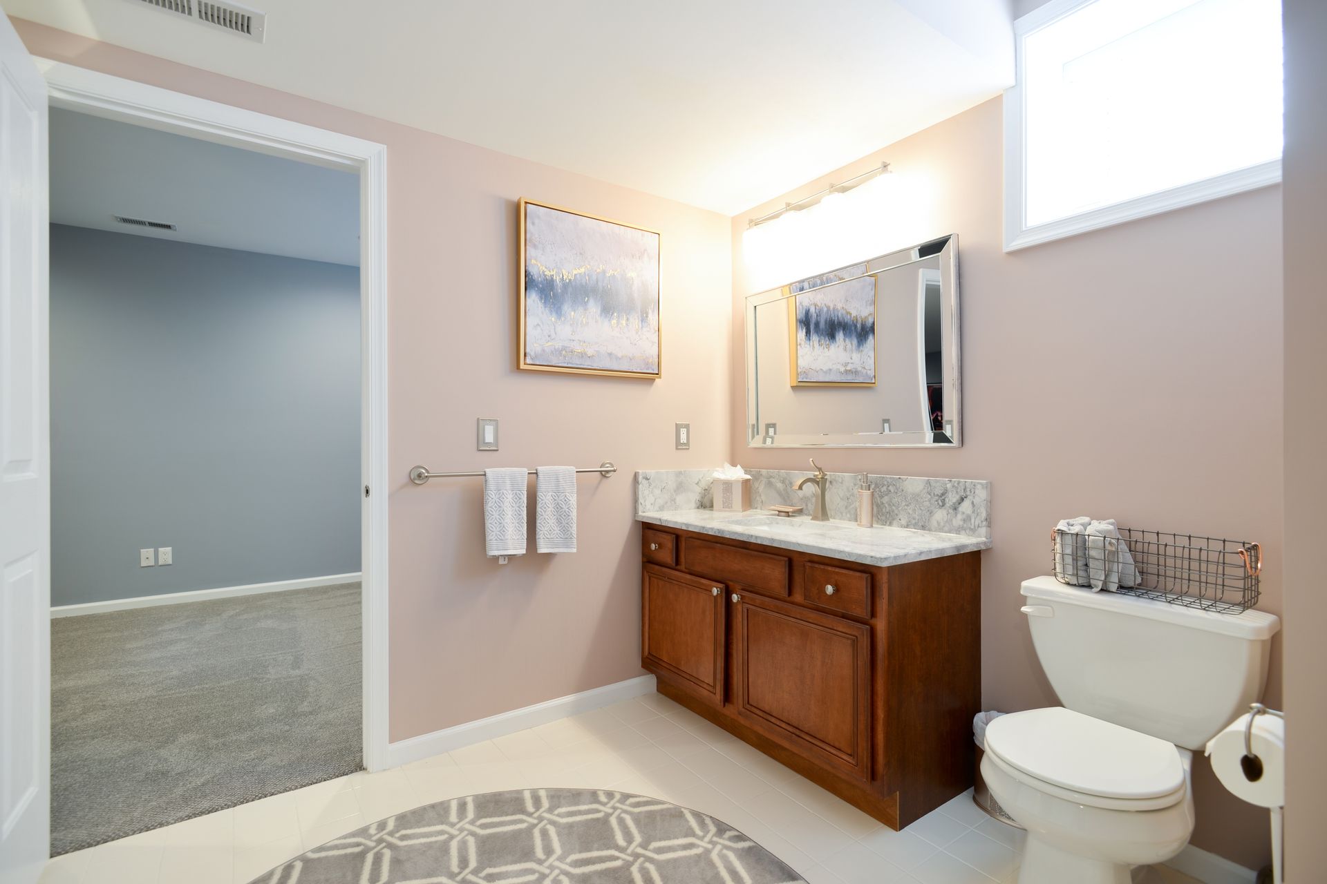 A bright bathroom with light pink walls, a wooden vanity, a large mirror, white toilet, and a doorway to a gray room.