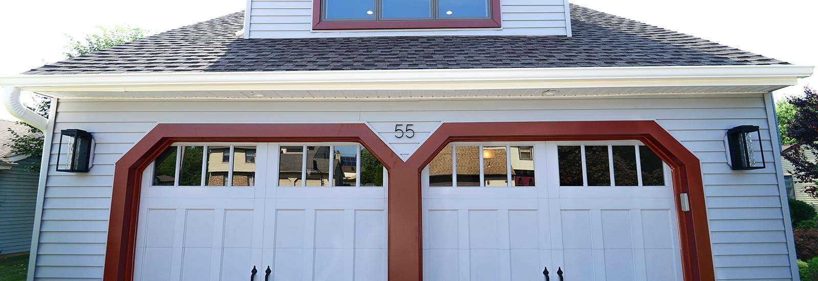 A white detached two-car garage with light gray siding, red trim around the doors, and a dormer window above.