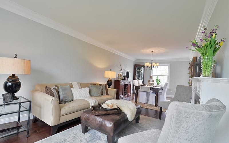 A living room featuring a tan sofa with throw pillows, a dark leather ottoman, and a dining area in the background.