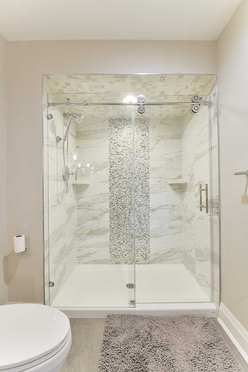 A modern walk-in shower with white marble-patterned tiles, a central mosaic accent strip, and glass sliding doors.