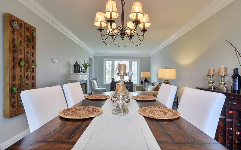 A dining room with a wooden table, white chairs, a decorative wall piece, and a chandelier, leading into a living area.