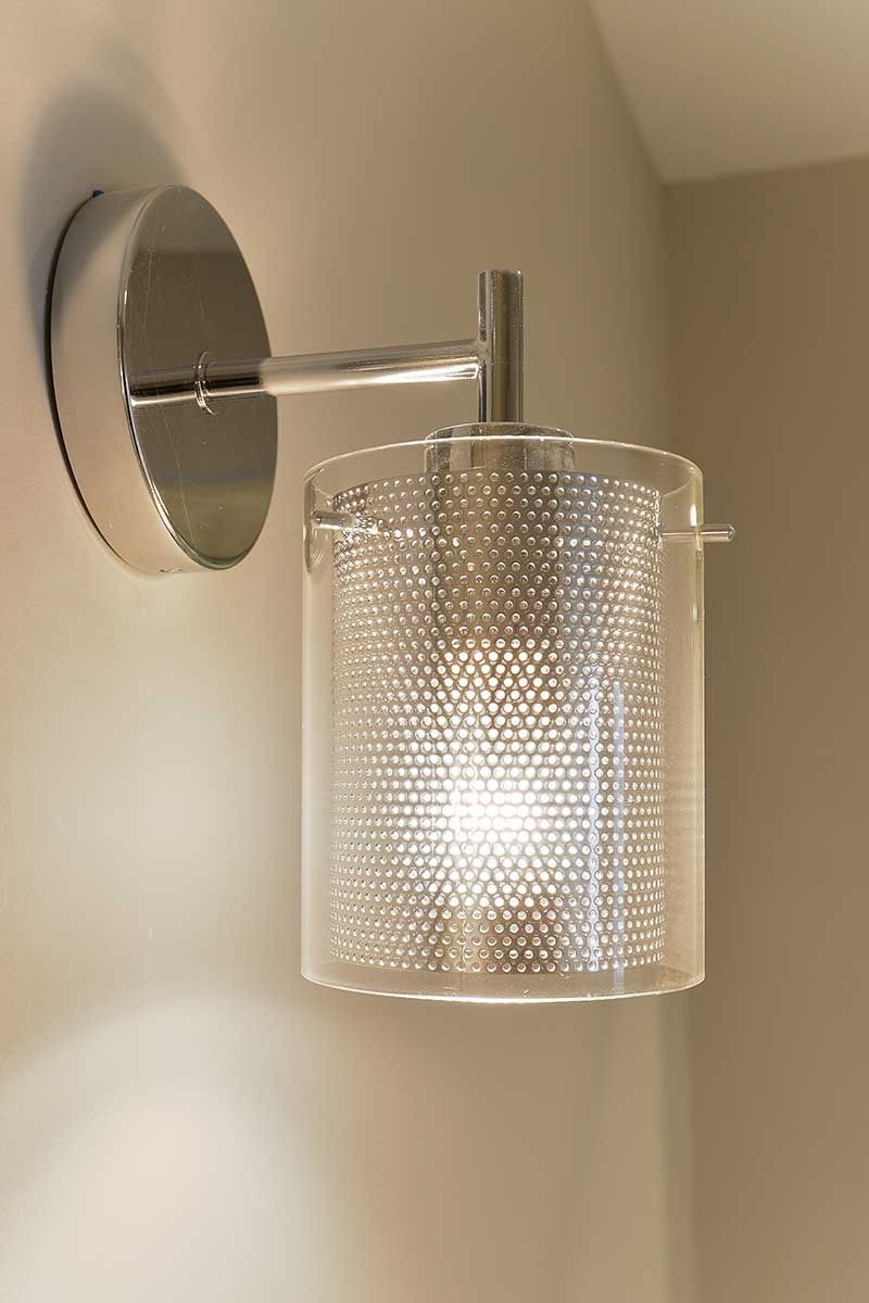A wall-mounted light fixture featuring a chrome finish and a cylindrical shade with a fine, perforated metal pattern.