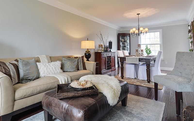 Living room with a beige sofa, dark leather ottoman with a faux fur throw, and an adjacent dining area with a chandelier.