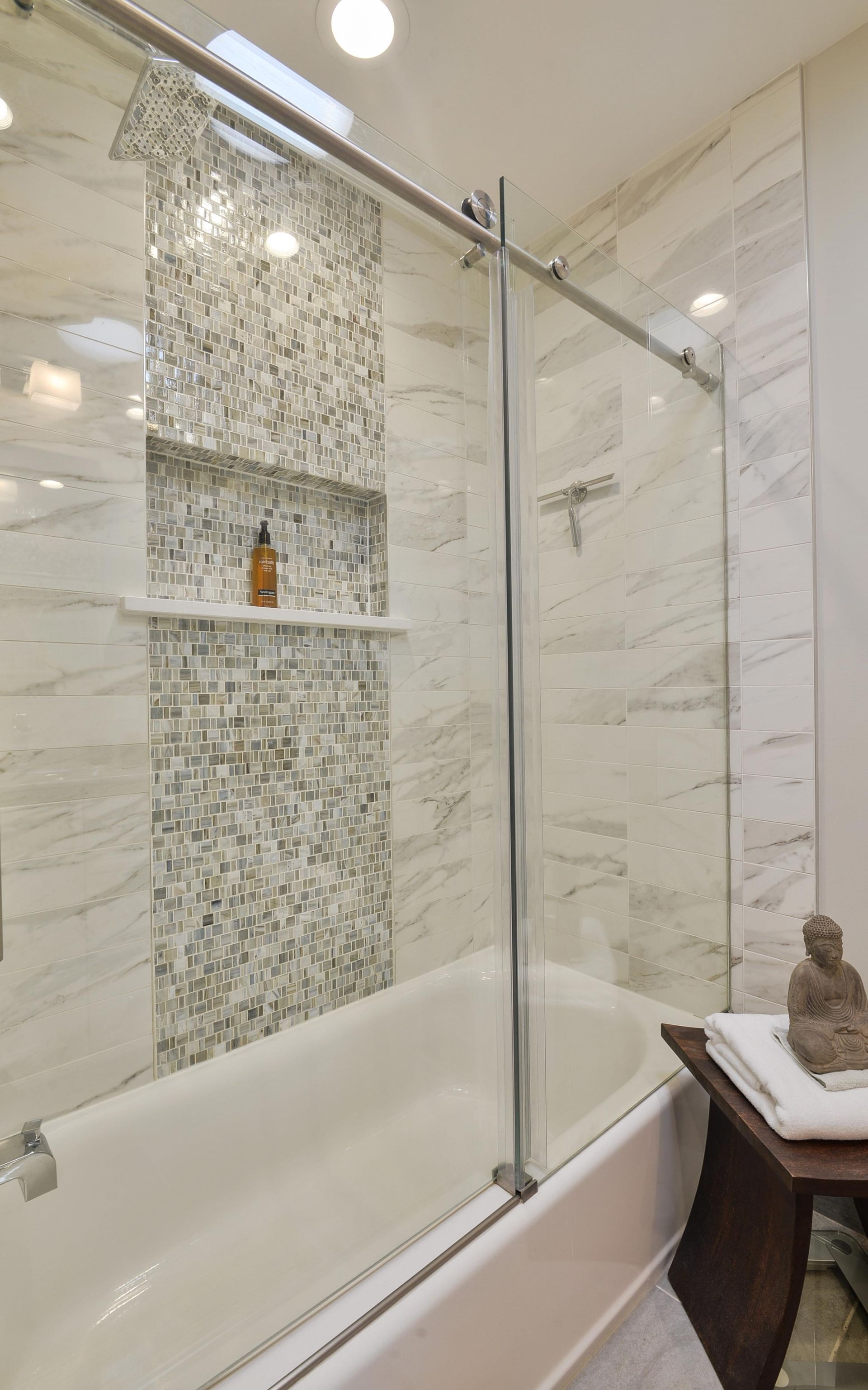 A modern bathroom featuring a white bathtub, marbled walls, a tiled shower niche, and a glass sliding door.