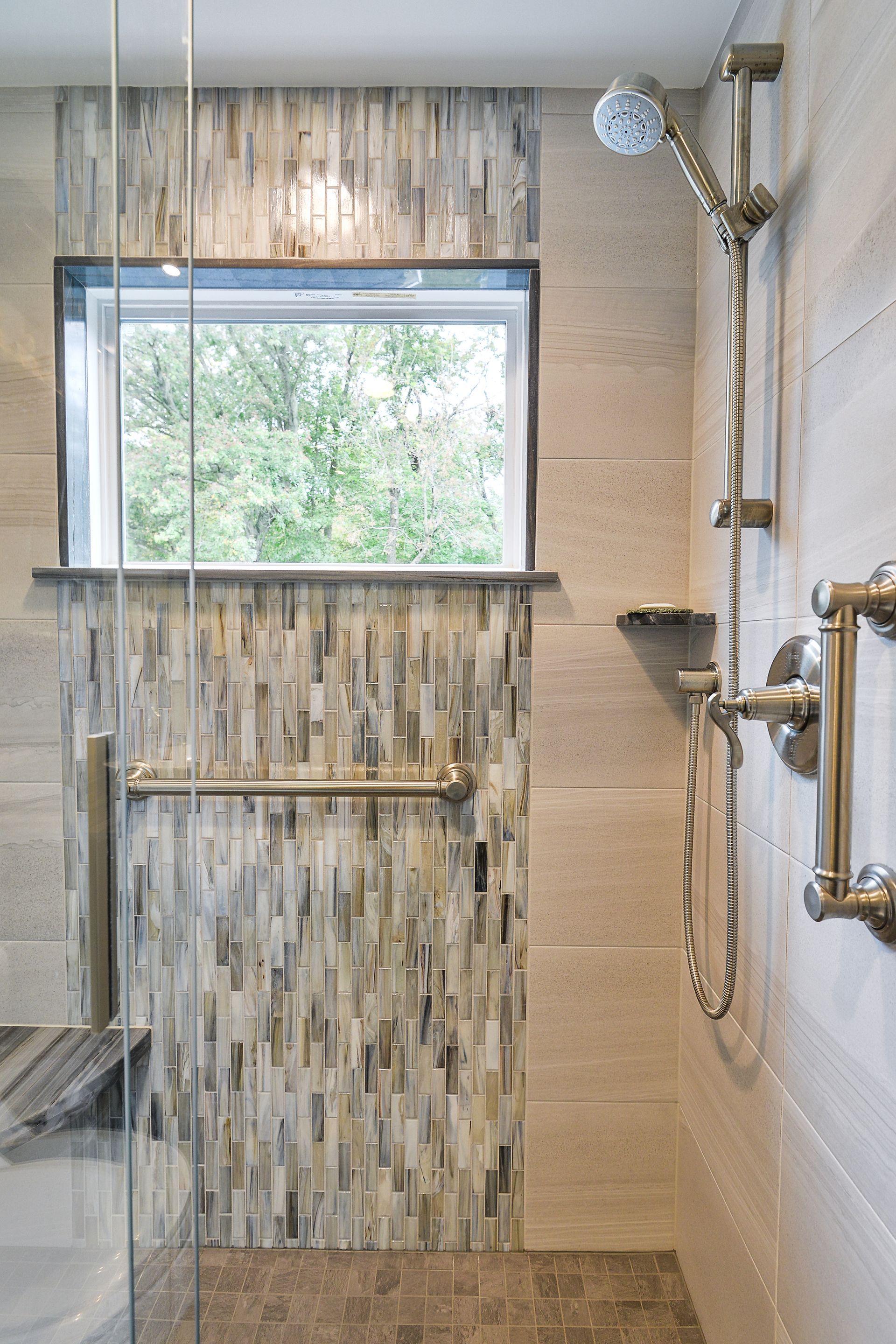 Modern shower with textured tile, a window, vertical stone tiles, and brushed gold grab bars and fixtures.