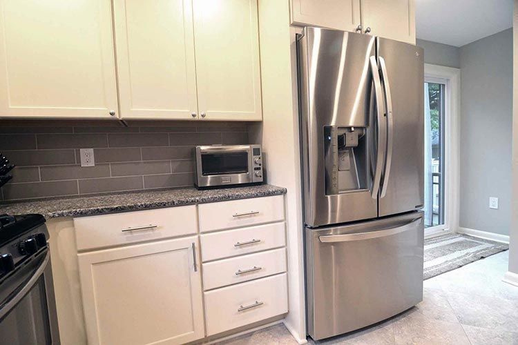 A modern kitchen featuring white cabinets, a gray tiled backsplash, granite countertops, a microwave, and a stainless fridge.