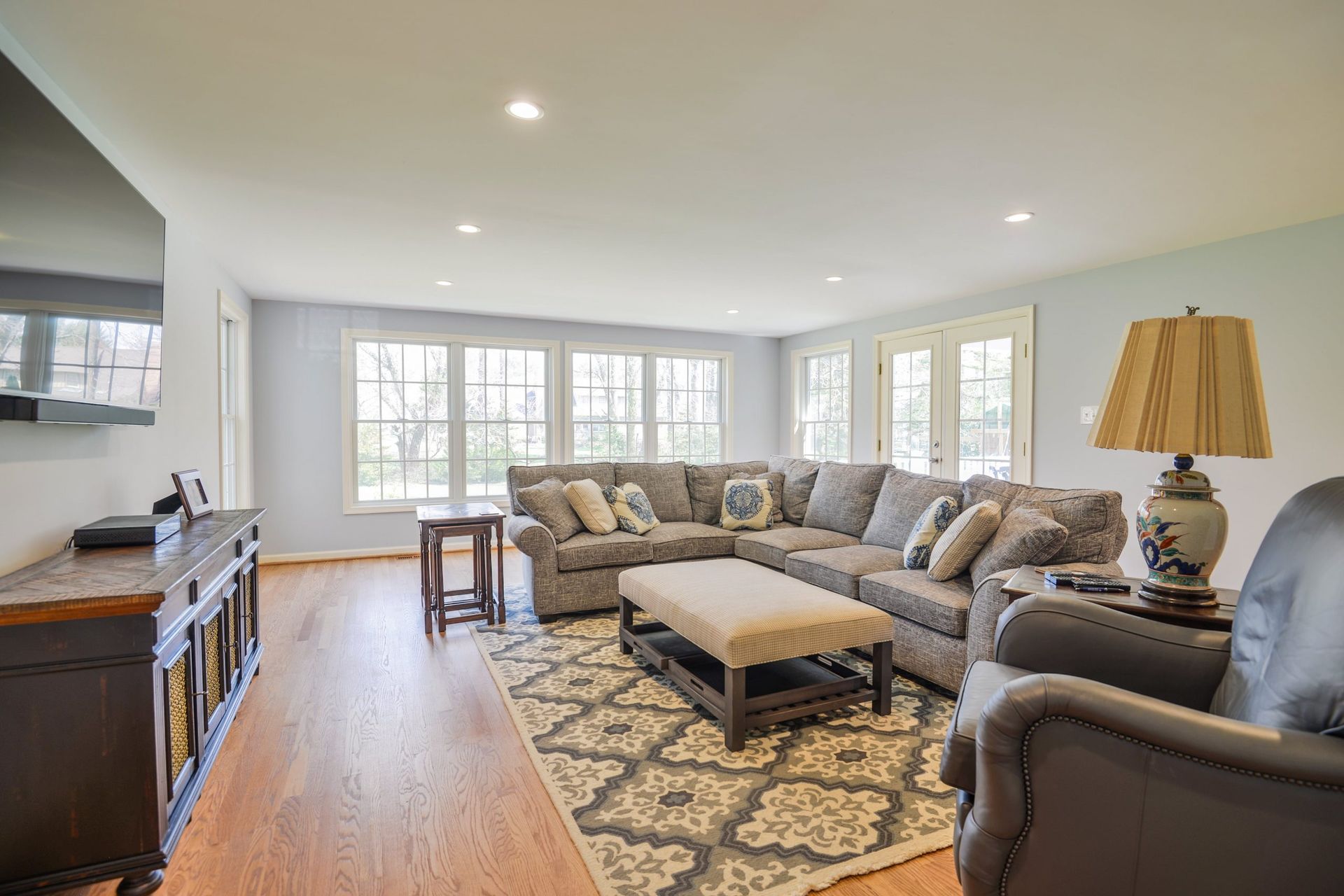 Living room with a large gray sectional sofa, patterned area rug, wooden furniture, and bright, sunlit windows.
