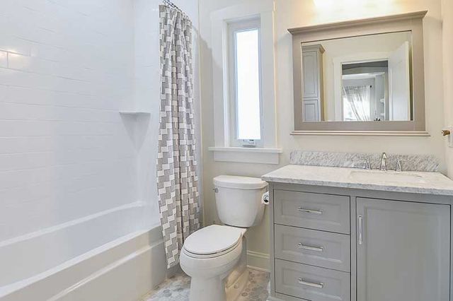 A bright, modern bathroom with a white bathtub, a grey vanity with a mirror, a window, and a white toilet.