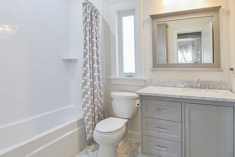 A bright, modern bathroom with a white bathtub, a grey vanity with a mirror, a window, and a white toilet.