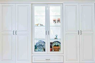 A white, symmetrical storage unit with two tall side cabinets flanking a central glass-fronted display cabinet and drawer.