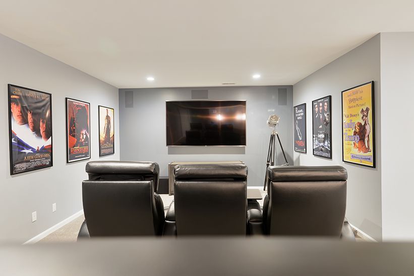A home theater room with three dark leather recliners facing a wall-mounted television, framed posters on gray walls.