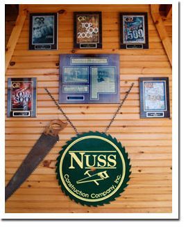 A wooden wall displays a circular Nuss Construction Company sign, a hanging saw, and several framed industry awards.
