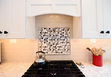 A kitchen stove with a black kettle and utensils in a red holder, featuring white cabinets and a gray hexagonal backsplash.