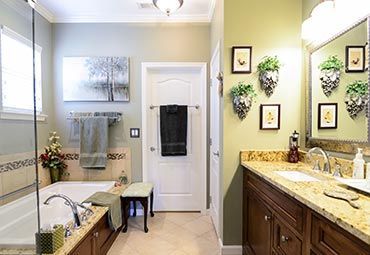 A bathroom featuring a beige bathtub, a wood vanity with a granite countertop, and sage green walls decorated with art.