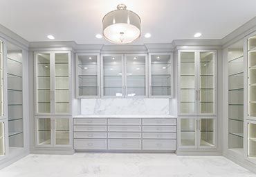 A symmetrical walk-in closet featuring light gray custom cabinetry, glass-front shelves, a marble backsplash, and drawers.