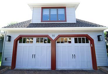 A two-car garage with white doors, red frames, grey siding, and a second-story window under a gabled roof.