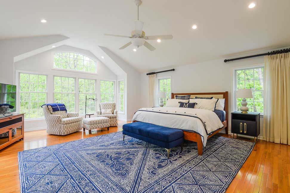 A spacious, bright bedroom with a king bed, blue patterned rug, armchair, and large windows with a view of greenery.
