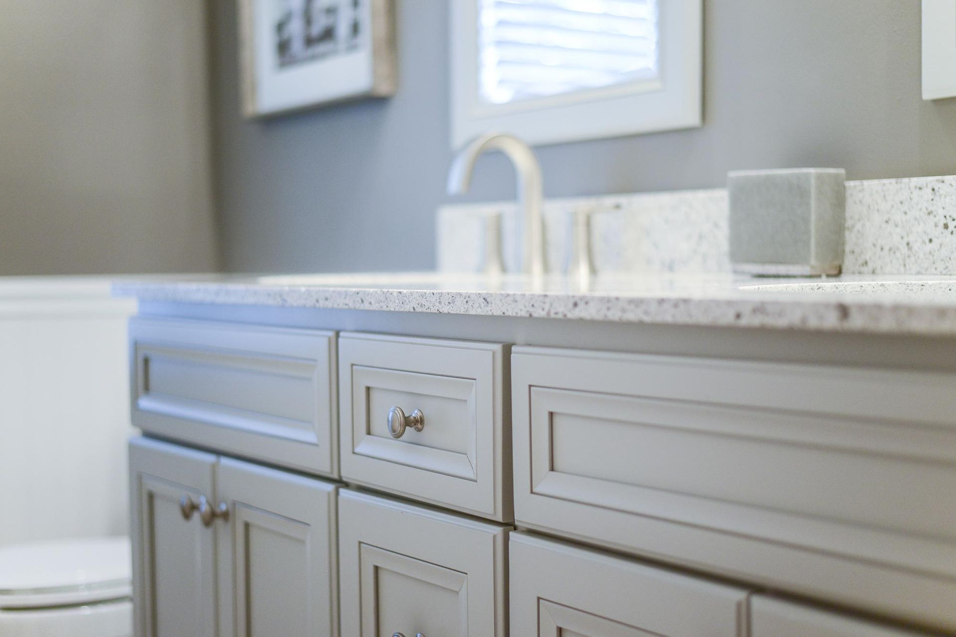 A light gray bathroom vanity with a speckled white countertop, silver faucet, and a small speaker on the counter.