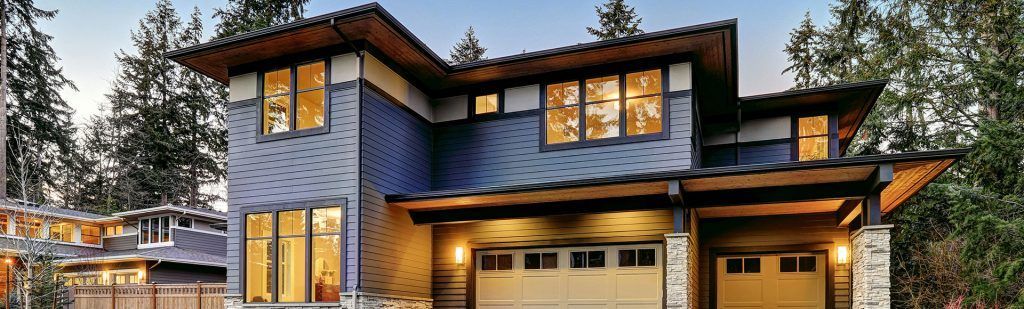Two-story craftsman-style house with blue siding, tan trim, warm lighting, and a multi-car garage in a wooded setting.