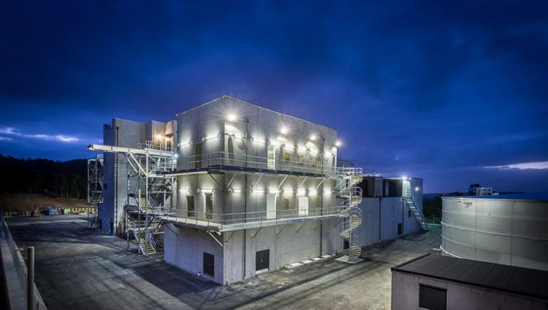 Exterior of a multi-level industrial building at dusk, lit with interior lights.