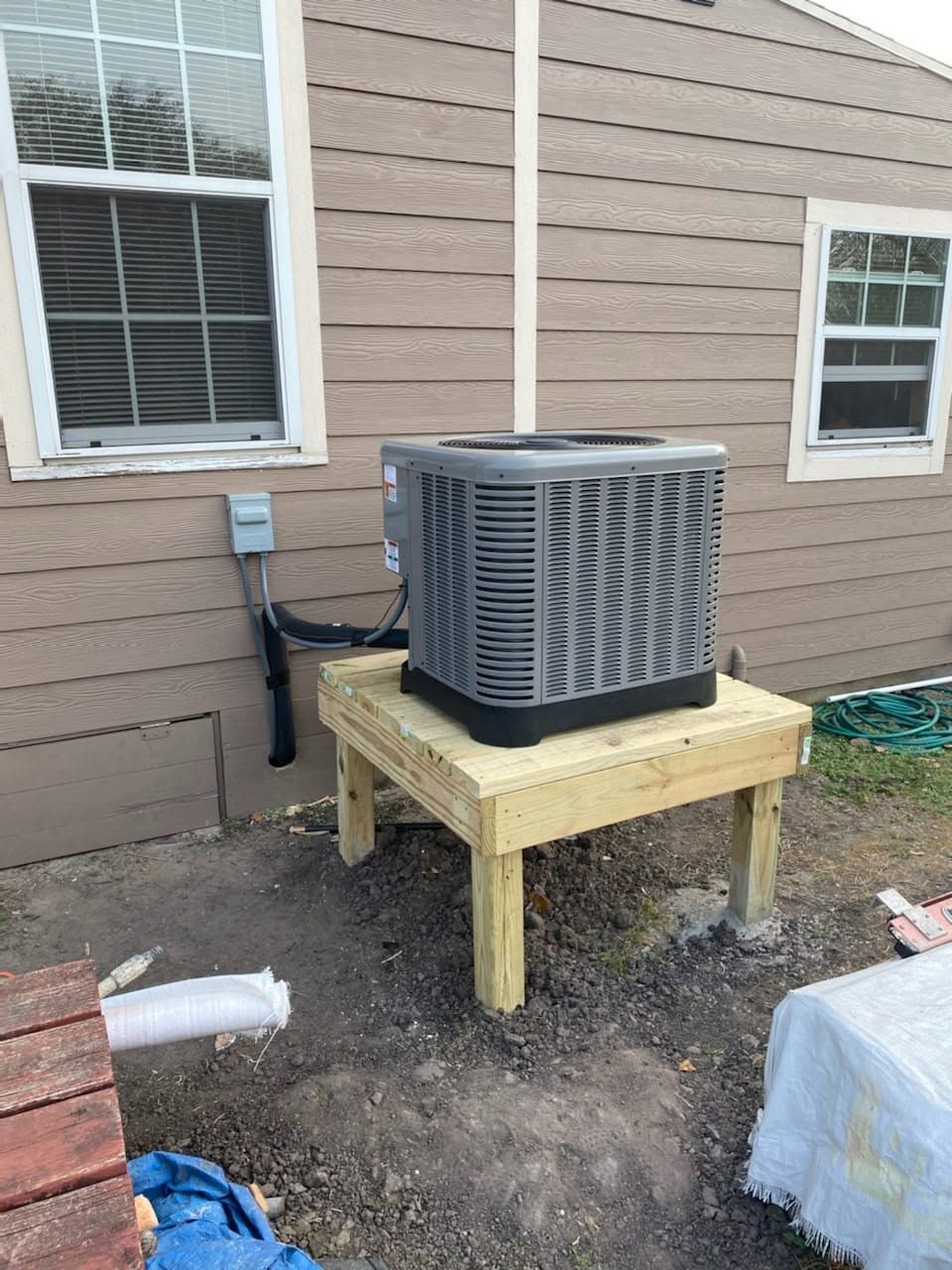Outdoor air conditioner unit on a wooden platform beside a beige house wall with windows.
