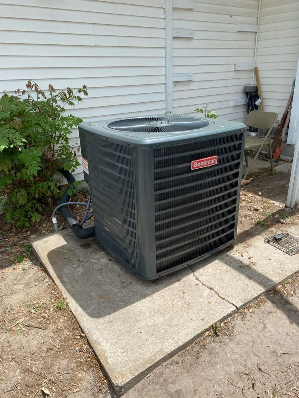 Outdoor central air conditioning unit beside a house on a concrete pad, with a chair and garden hose nearby.