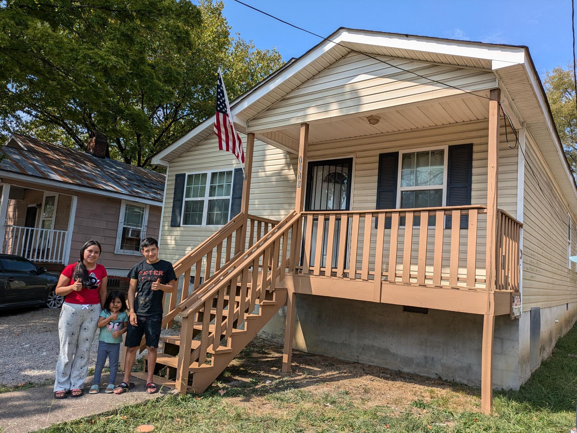 Family stands in front of a small house with an American flag. The house is tan with a porch.