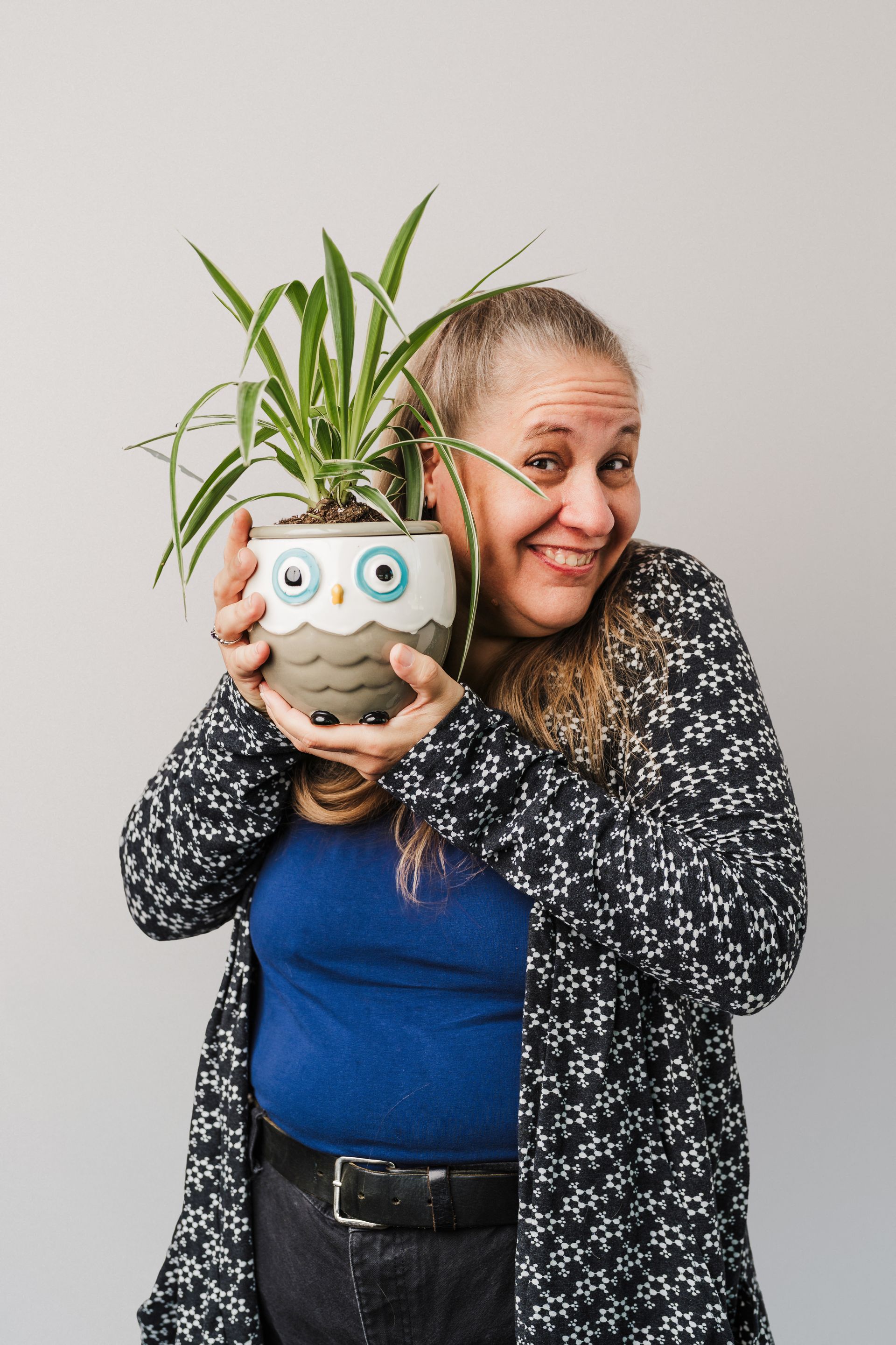 Woman holding owl-shaped planter with spider plant, smiling, wearing cardigan and blue shirt.