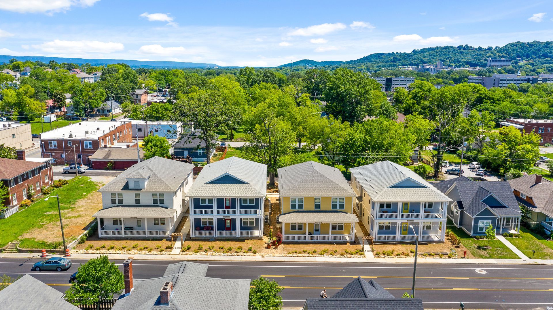 Row of four multi-story houses with front porches, trees, and distant hills.