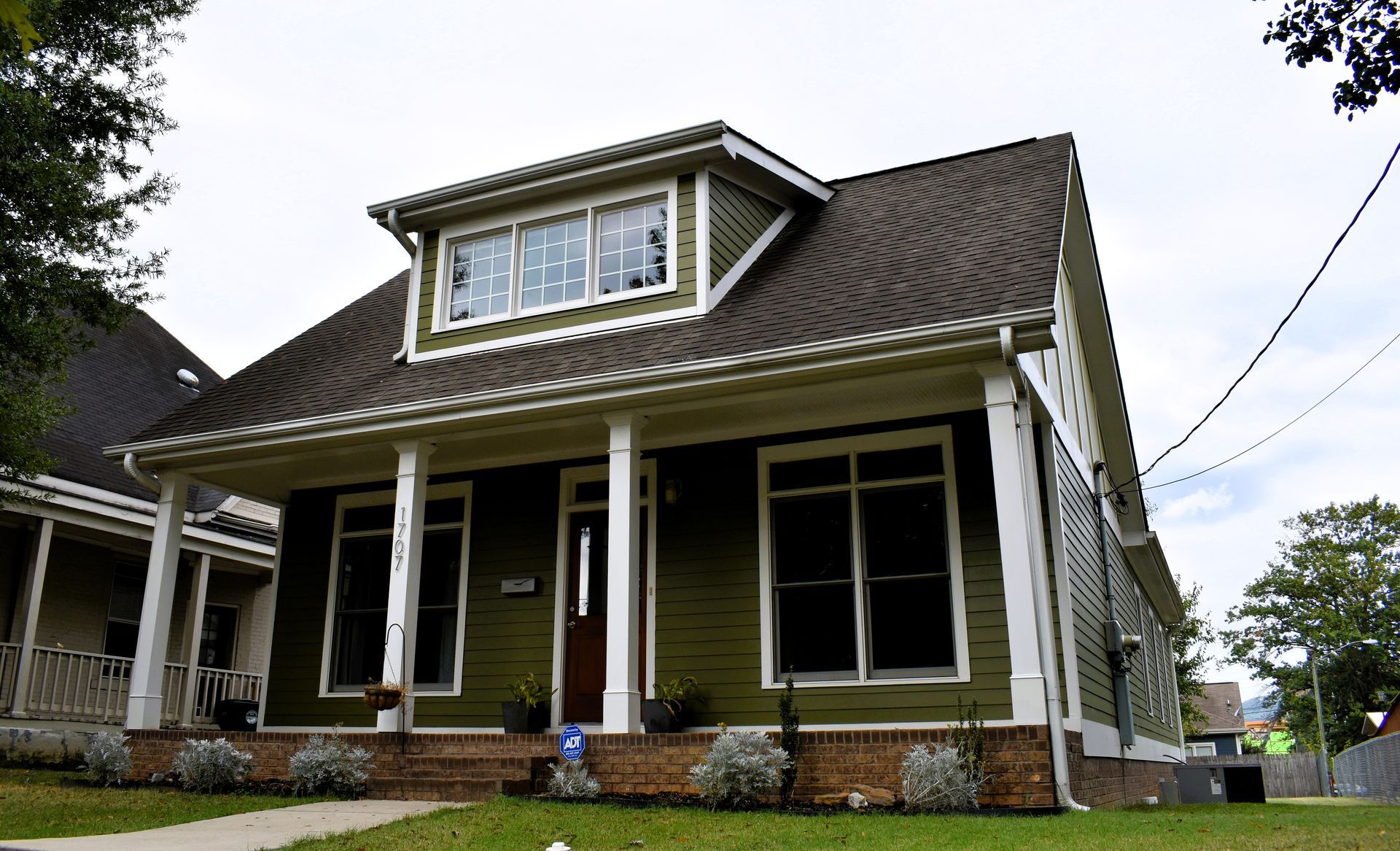 Green house with white trim, porch, and dormer under a cloudy sky.