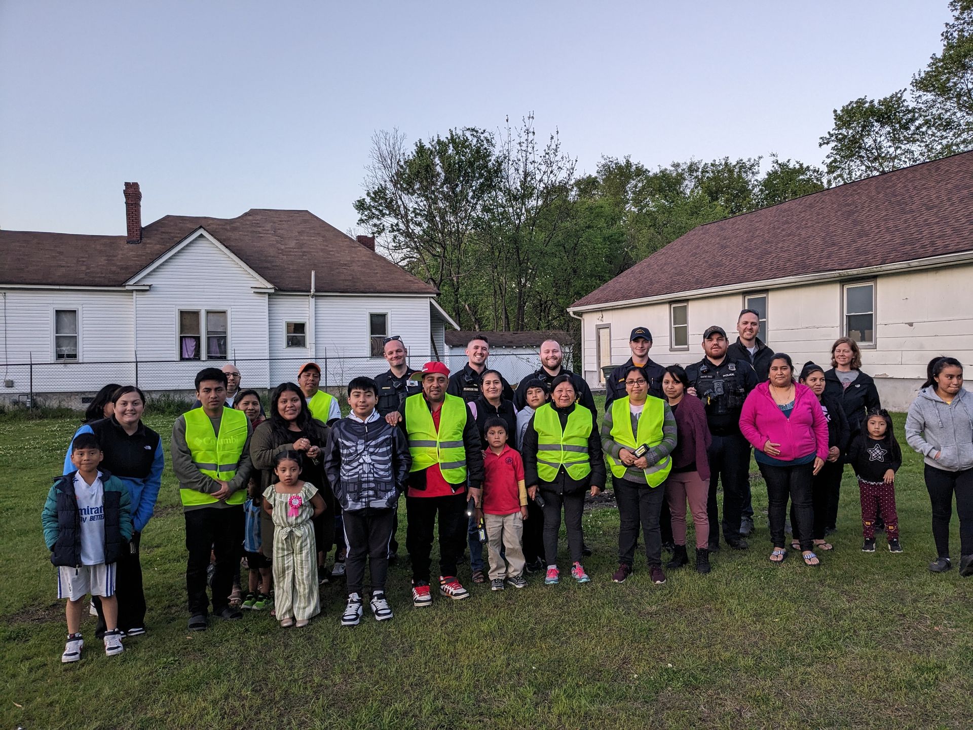 Group of people, some wearing yellow vests, standing outside two houses on a grassy lot.