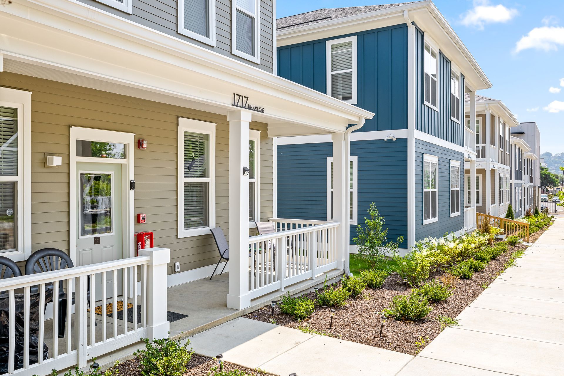 Multi-story townhomes with front porches and landscaping, featuring tan, gray, and blue siding, with a sidewalk running along the front.