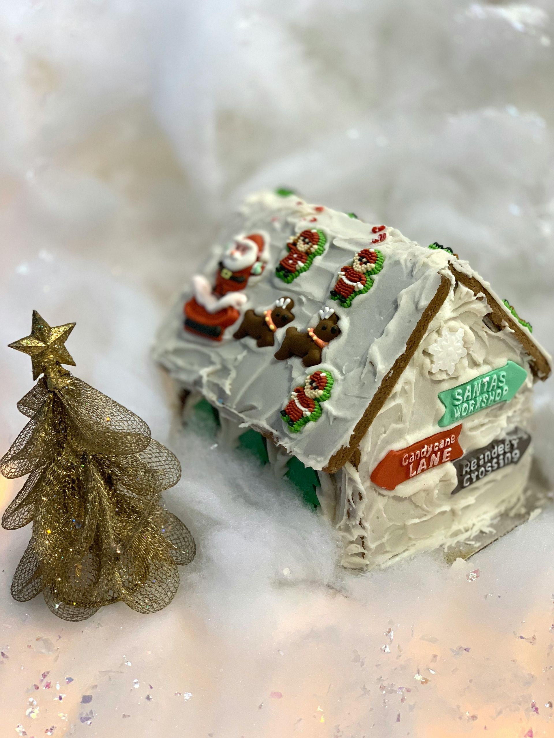Gingerbread house decorated with candy in snowy setting, next to a gold Christmas tree.