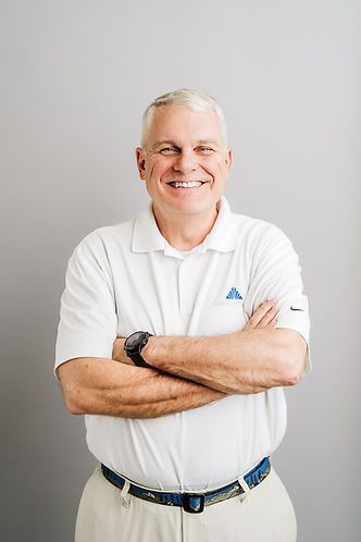 Smiling man in a white polo shirt and khaki pants with arms crossed against a grey background.