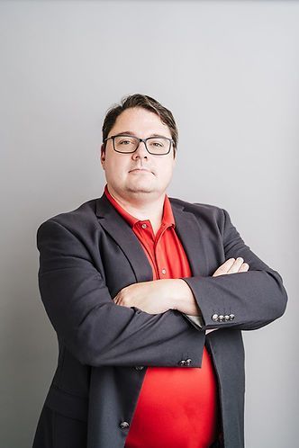Man with glasses, arms crossed, wearing a red shirt under a dark blazer, against a gray background.