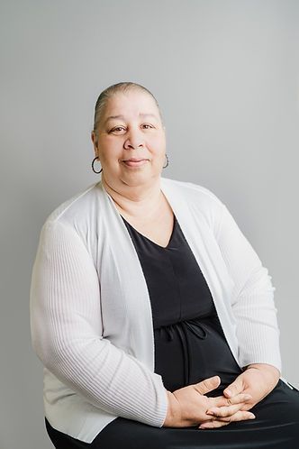 A Black woman with a shaved head, wearing a black dress and white cardigan, smiles at the camera. Gray background.