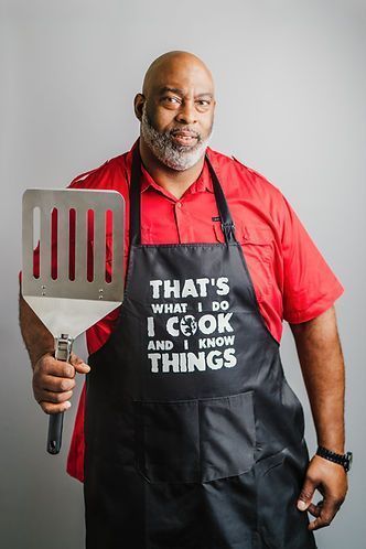 Man wearing a red shirt and apron, holding a spatula. Apron says, 