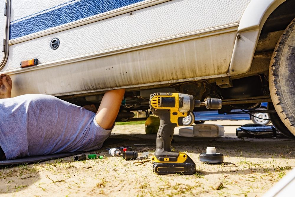 Person lying under RV, using power drill for repairs; outdoors. — The Caravan Clinic in Tweed Heads, NSW
