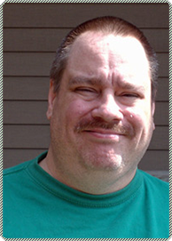 Smiling man in a green shirt standing outdoors against a brown wall