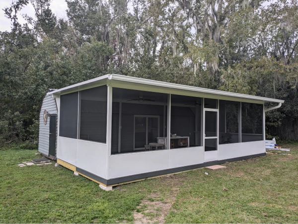 A white screened-in porch with dark screens and a gray roof, set in a grassy backyard. A small shed is attached to the side.