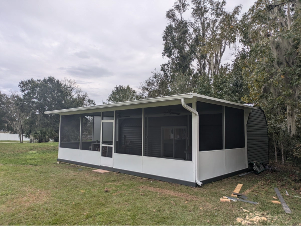 A white screened-in porch with a gray roof sits on a grassy yard, surrounded by trees under a cloudy sky.
