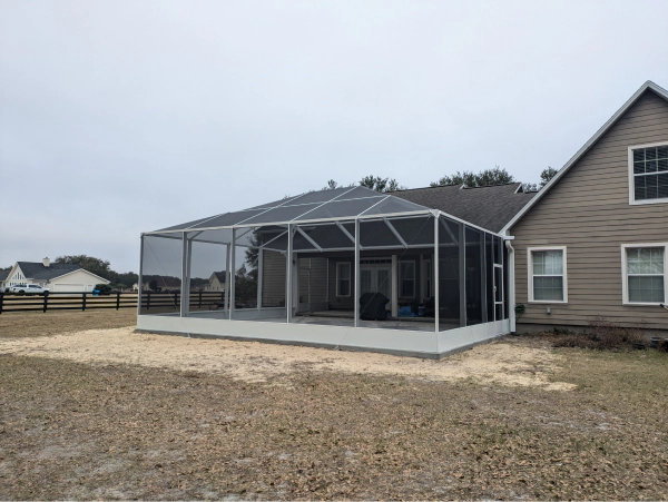 Screened-in patio attached to a beige house, on a gravel and grassy lot under an overcast sky.