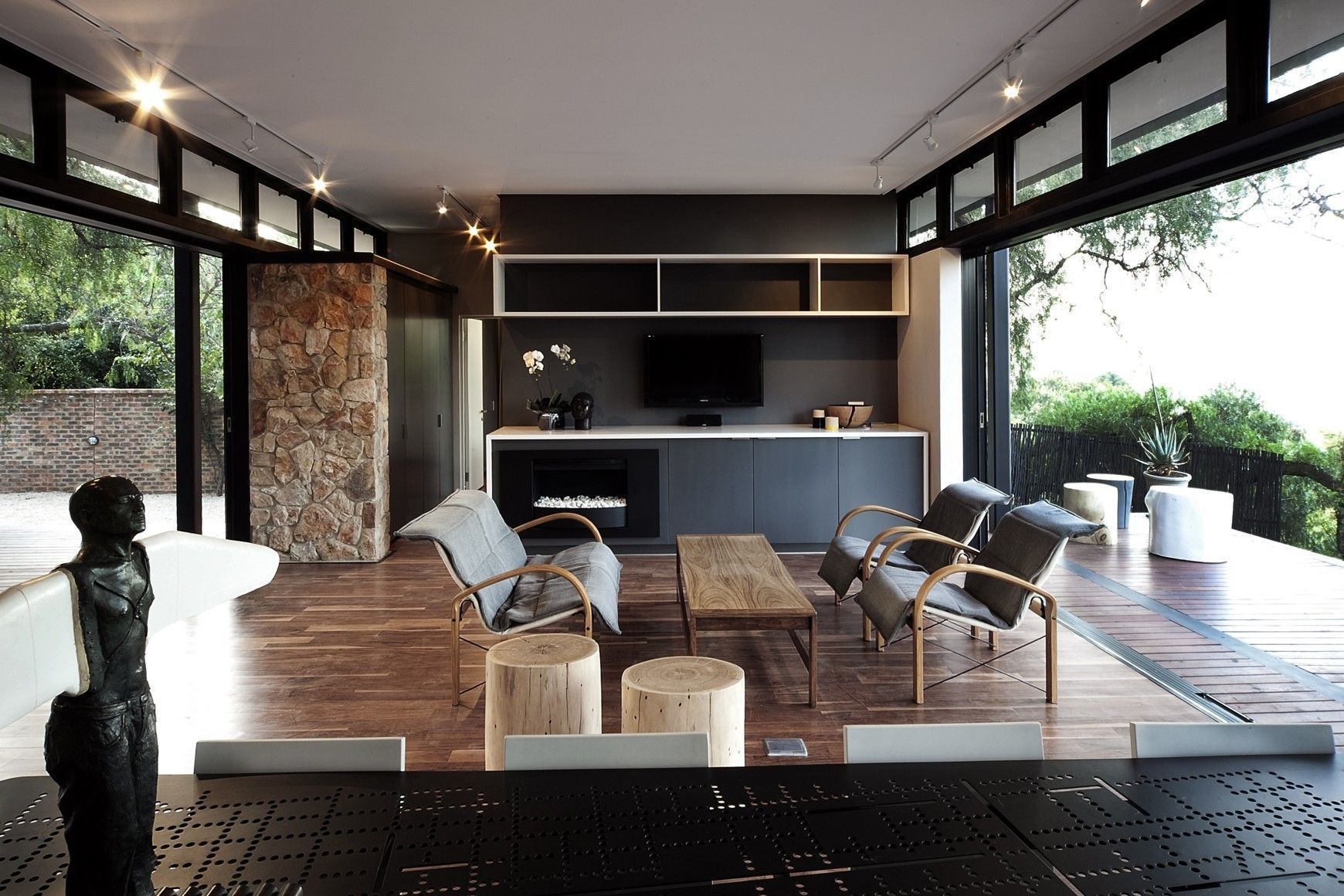 Salon moderne avec armoires gris foncé, mur en pierre, murs en verre ouverts et meubles en bois.