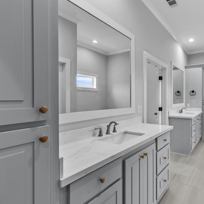 Modern double-sink bathroom with gray cabinetry, white countertops, and large mirrors along a bright hallway.